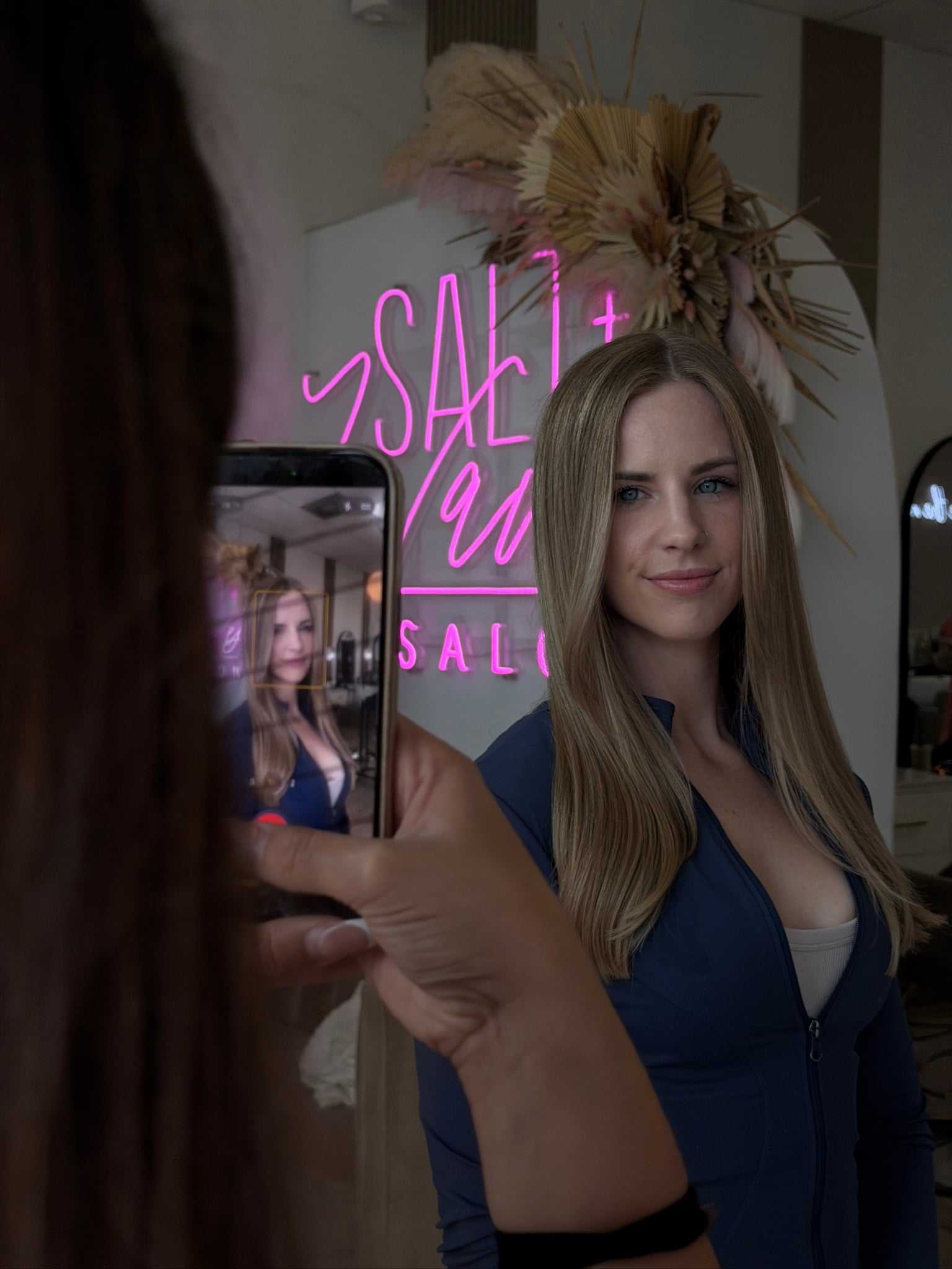 Woman posing in a salon, photographed with a smartphone, neon "salon" sign in background.