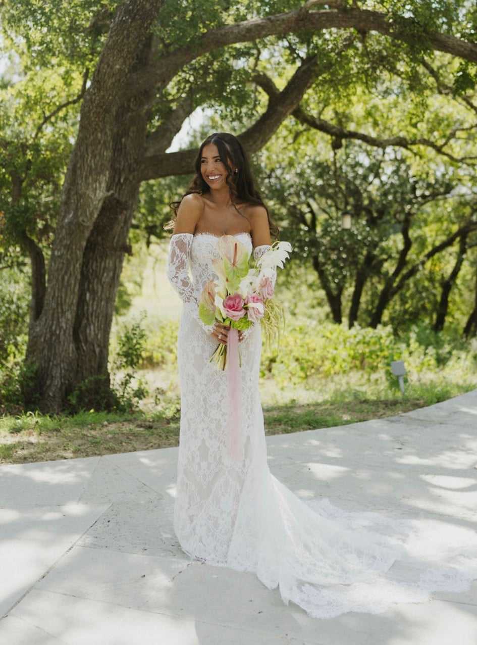 Bride in lace gown holding bouquet, standing outdoors with trees in the background.