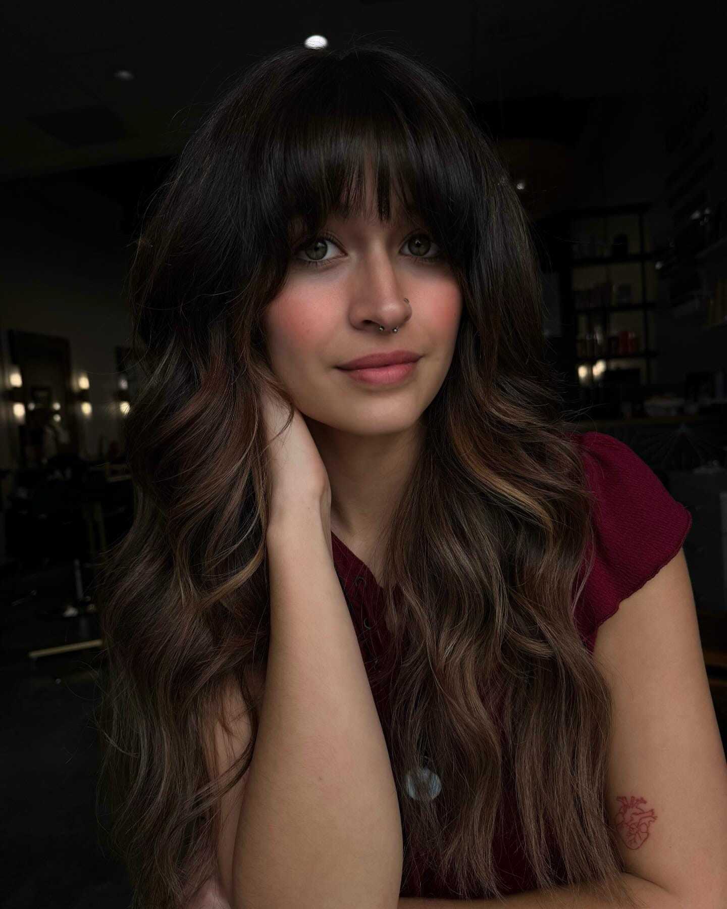A woman with long, wavy hair and a nose ring sits in a dimly lit salon, wearing a burgundy top.