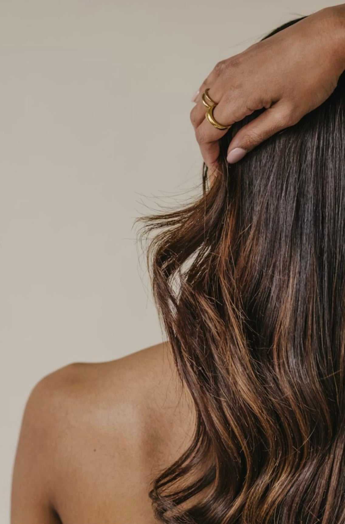 Hand in gold rings touching wavy, brown, shoulder-length hair on a bare shoulder.