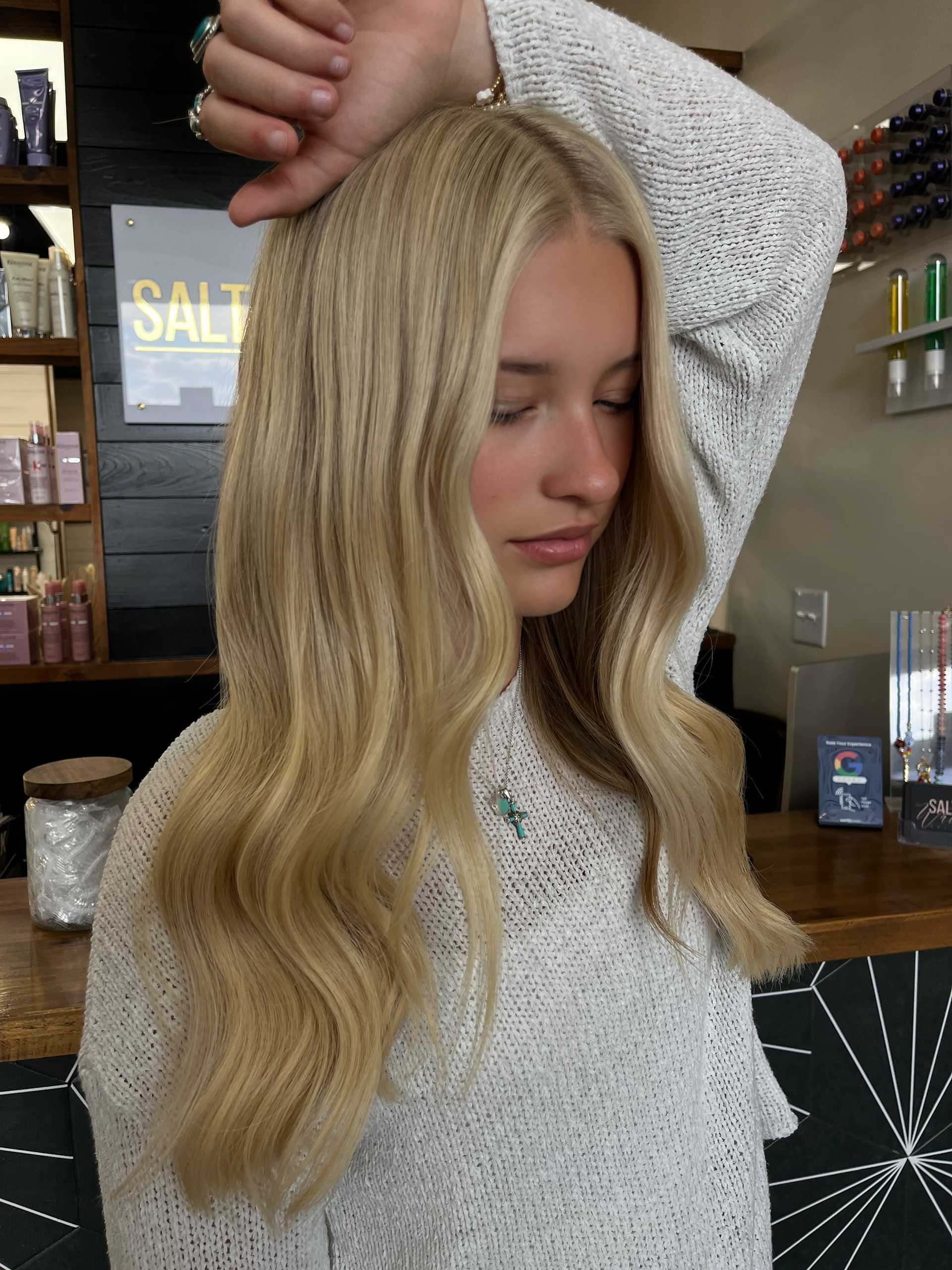 Blonde woman with long, wavy hair in a salon setting, wearing a white sweater.